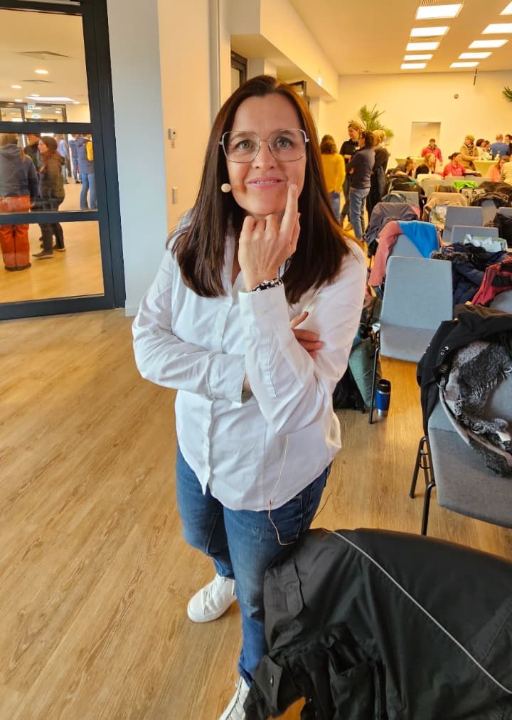 Eine Frau mit Headset-Mikrofon steht lächelnd in einem Seminarraum, im Hintergrund Teilnehmende zwischen Stühlen und Jacken.