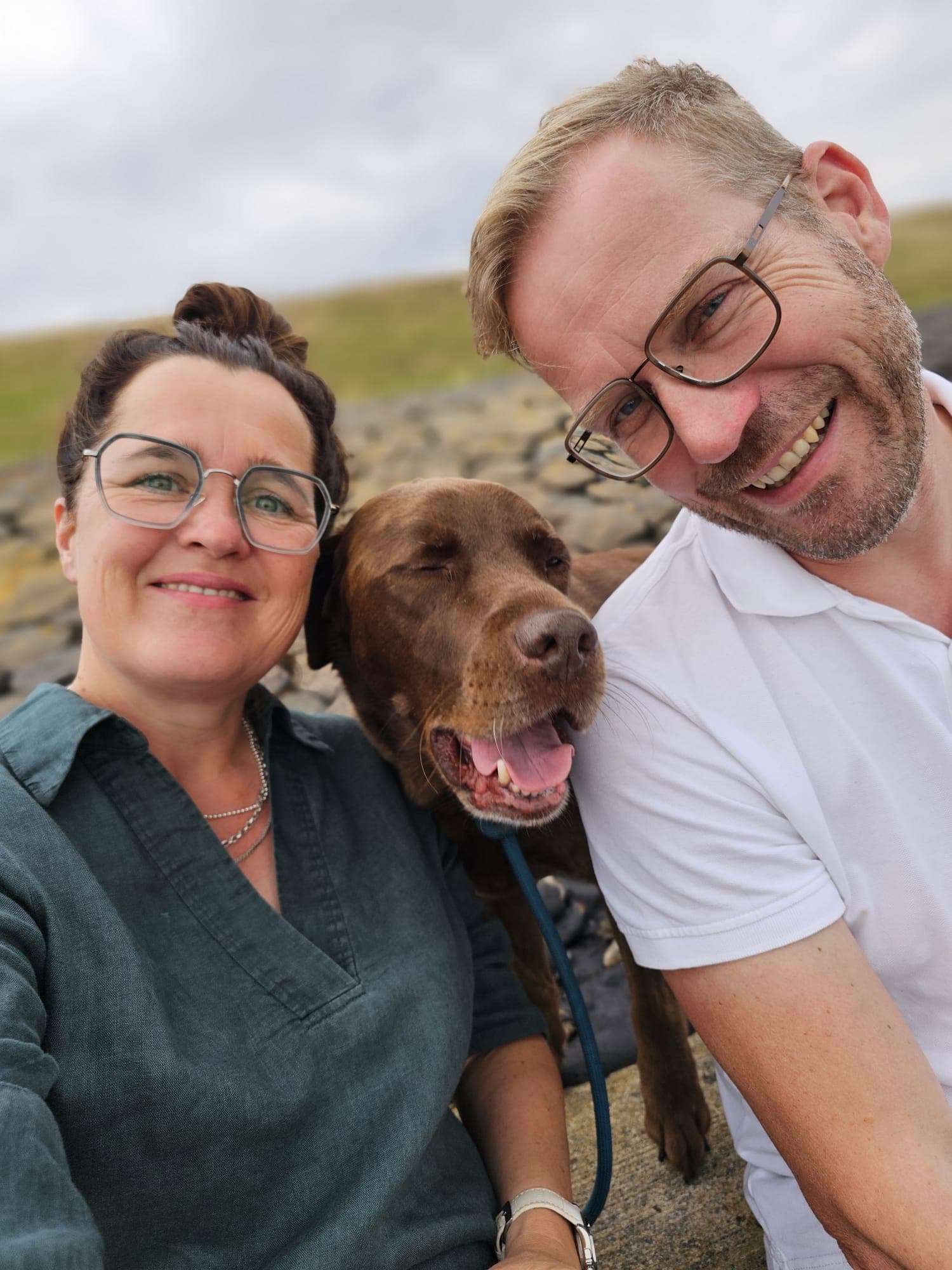 Raphaela Niewerth mit ihrem Mann und ihrem Schokoladen-Labrador an der Kueste, alle drei lachen in die Kamera.